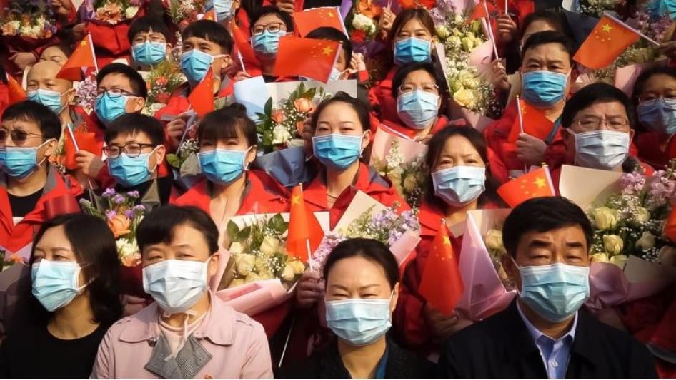 Image of people wearing face masks and holding Chinese flags and brightly-colored flowers.