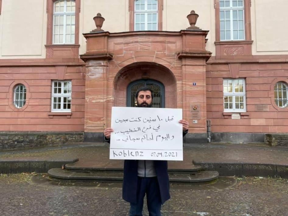عامر مطر يحمل لافتة مكتوب عليها بالعربية، "قبل 10 سنين كنت سجين في فرع الخطيب... واليوم أحاكم سجاني...".