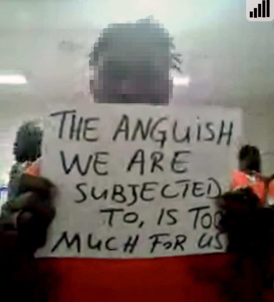 A man with a blurred face holds up a sign from inside a detention center