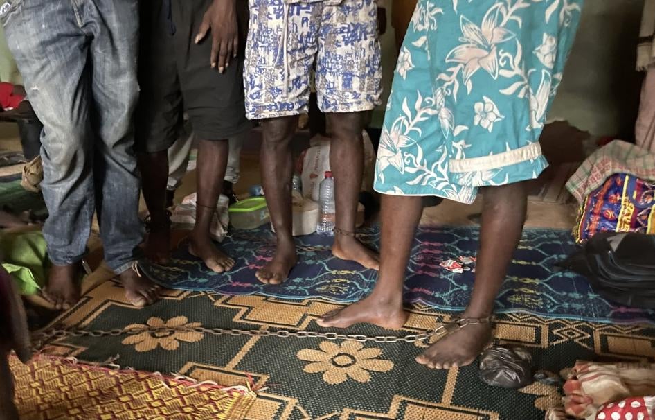 There are four people standing, all of whom have their ankles chained to restrict their movement.
