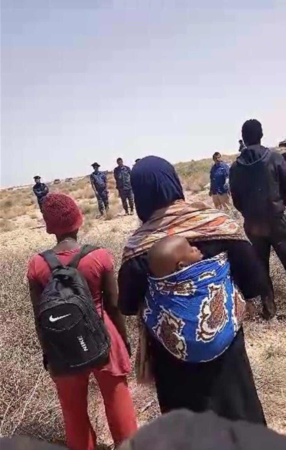 Un groupe de migrant·e·s et de demandeur·euse·s d’asile noir·e·s de plusieurs nationalités africaines, dont une femme et son bébé, bloqué·e·s dans le désert depuis des jours après avoir été expulsé·e·s de Tunisie, debout dans la zone tampon à la frontière entre la Tunisie et la Libye, face à une équipe de journalistes d’Al Jazeera et à des soldats libyens, le 11 juillet 2023.  © 2023 Privé