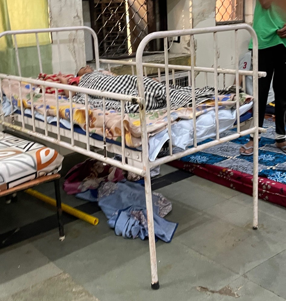 An individual lying in a metal-framed bed inside the Asha Kiran institution.