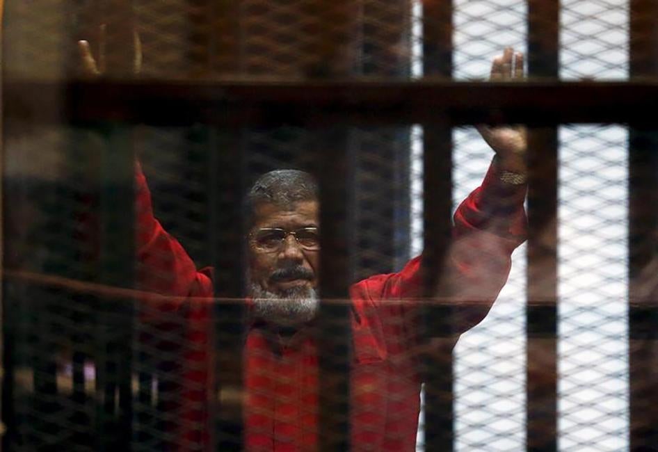 الرئيس المعزول محمد مرسي في البذلة الحمراء، الزي الخاص بالمحكومين بالإعدام، يُحيّي محاميه وأشخاص آخرين من وراء القضبان أثناء ظهوره في المحكمة في يونيو/حزيران 2015 مع أعضاء آخرين من الإخوان المسلمين، في ضواحي القاهرة. 