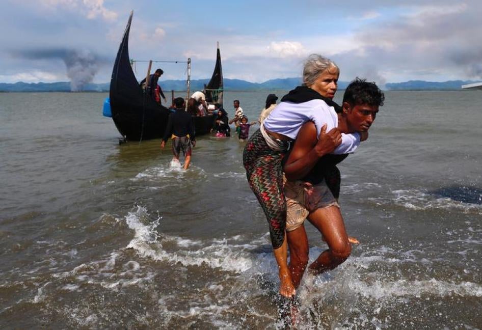 一名年老的罗兴亚女性难民乘船横渡孟加拉湾，跨越孟加拉与缅甸的海上边界之后，被人背上岸边，远方缅甸境内则仍烽烟四起，孟加拉，沙波里尔堆，2017年9月15日。