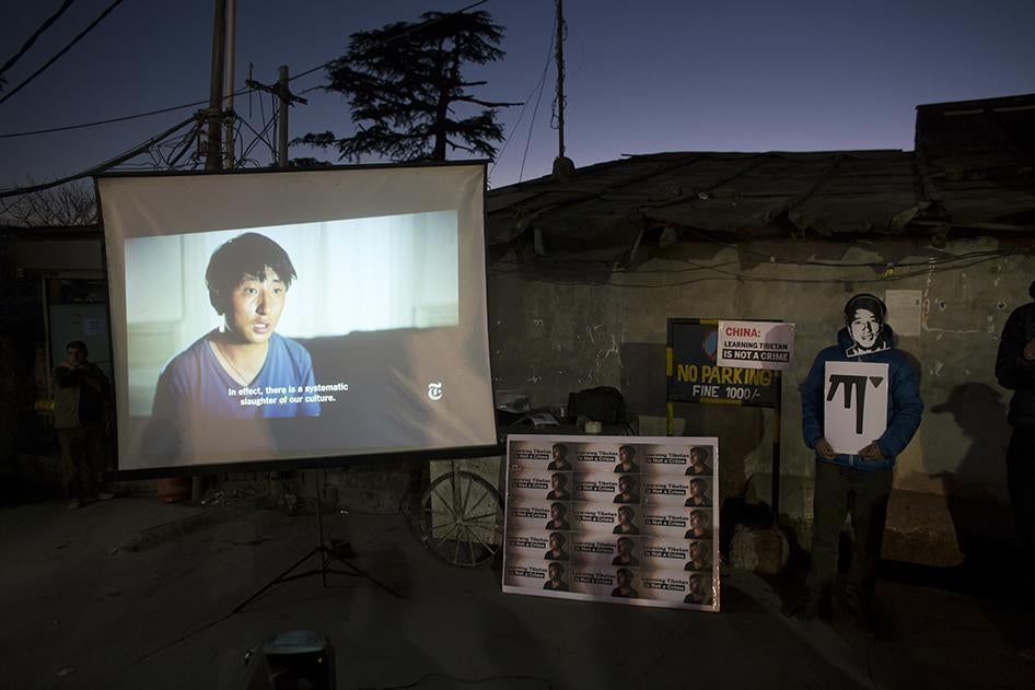 An exile Tibetan wearing a mask in the likeness of Tashi Wangchuk, stands next to a screen projecting a New York Times video during a street protest demanding his release, in Dharmsala, India. © 2018 AP Photo/Ashwini Bhatia