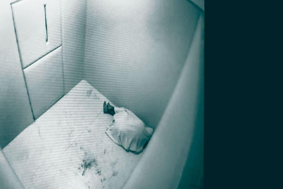 A person lying on the floor of a prison cell, seen through a CCTV camera.