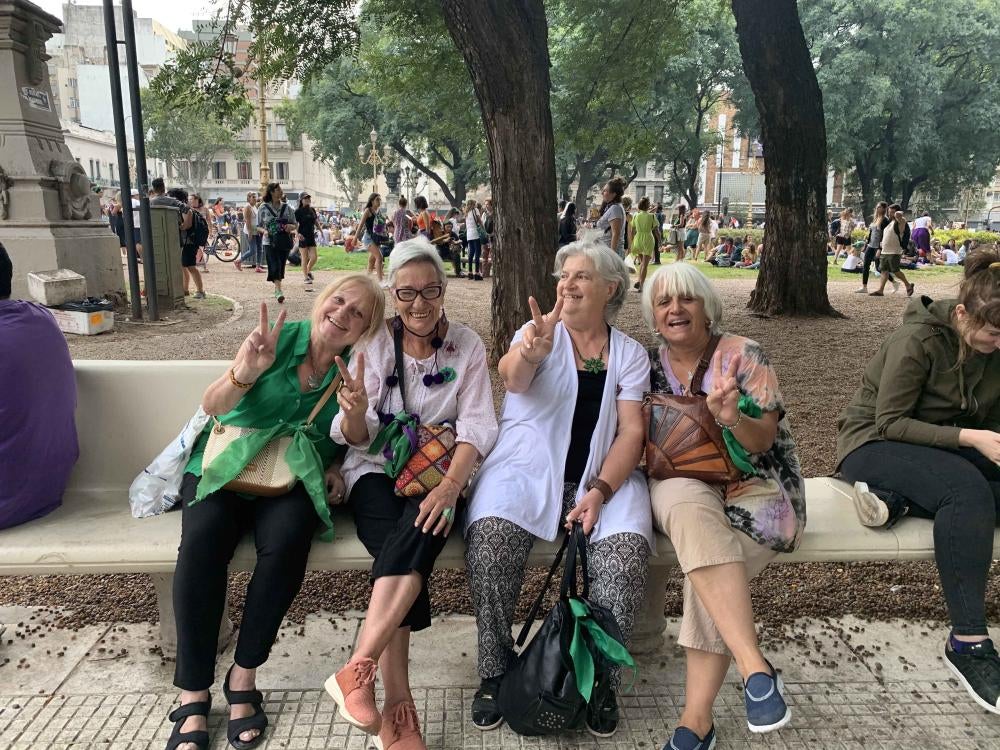 Four women hold up peace signs and pose for a photo at an outdoor rally