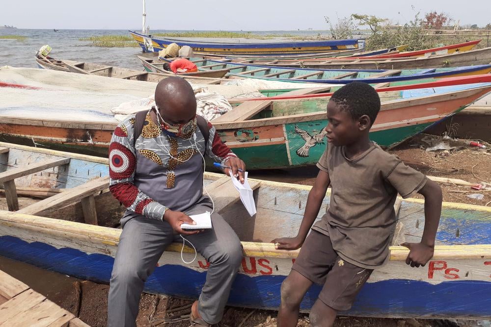 A man takes notes while interviewing a boy while sitting on a wooden boat on the beach