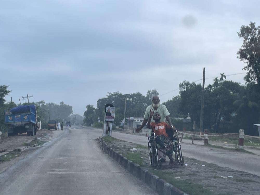 Un homme debout derrière son fils handicapé, assis dans un fauteuil roulant, mendiait dans une rue de Sylhet, une ville du Bangladesh qui a été inondée en juin 2022. Les inondations sans précédent dans le nord du pays ont causé des difficultés extrêmes, en particulier pour les personnes handicapées et leurs familles. 