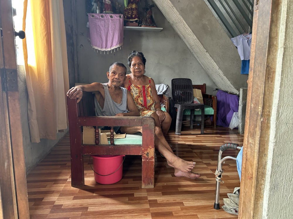 Aidaliza Aquino supports her husband, Eduardo Aquino, who uses a walker for mobility, at the Pulo relocation site in Barangay Candelaria, Macabebe, Pampanga, Philippines, July 13, 2026.