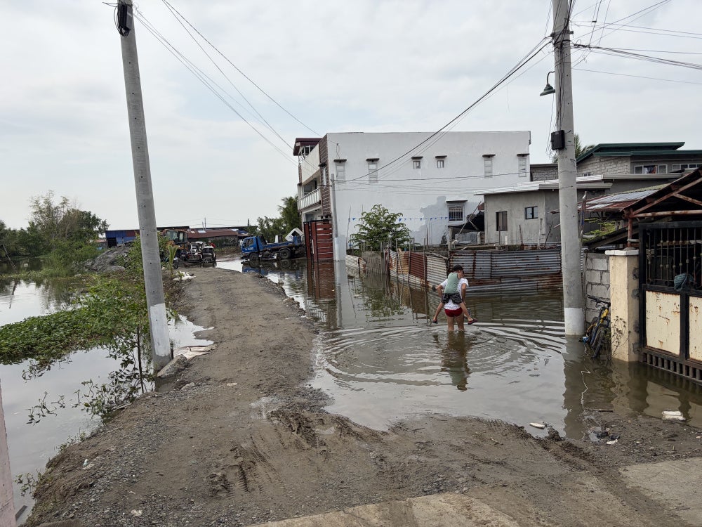 A man carries a boy on his back as they wade through floodwater around the Pulo relocation site, July 14, 2026.