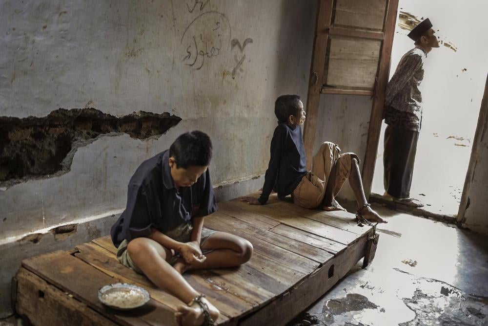 Dua penghuni di pusat pengobatan Bina Lestari Brebes, Jawa Tengah,dirantai pada permukaan kayu tempat tidur saat seorang penyembuhkeagamaan berdiri di dekatnya. Di tempat ini, semua penghunidirantai dan menerima “pengobatan” tradisional melalui doa,konsum