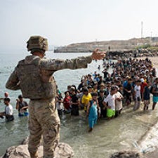 Figure in military garb addresses large group of migrants.