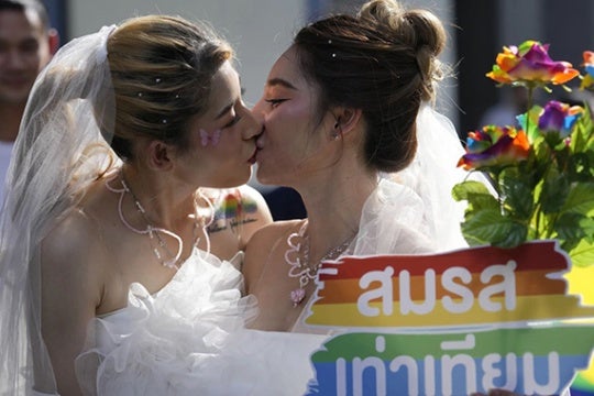Two women in wedding dresses kiss. 