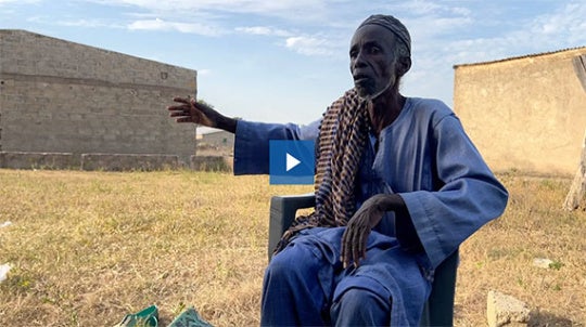 Figure sits in chair, facing camera. 