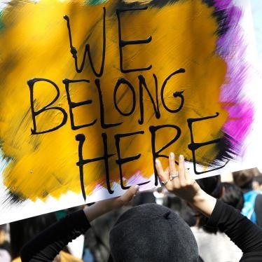 People demonstrate at a rally in Chinatown, New York City. Someone is pictured holding a painted sign reading "We Belong Here" with yellow, blue, pink, and black colors..