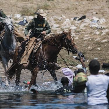 Two Customs and Border Protection agents on horseback approach three Haitian migrants standing in the river. The migrants are holding bags and shoes.