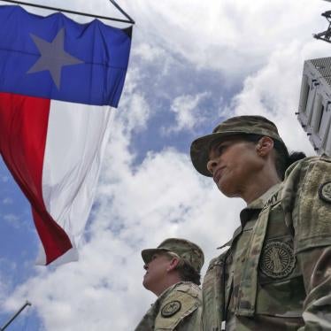 Low angle photograph looking up at a stern faced Army National Guard solider who stares ahead. The Texas flag flutters overhead.