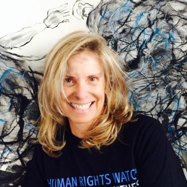 A woman with shoulder length blonde hair is wearing a black t-shirt that says "Human Rights Watch" in blue text and "Tyranny has a witness" in white text. She is smiling and standing in front of a black and blue painting.