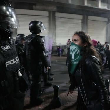 A woman wearing a green bandana over her face stands in front of a police officer with riot gear