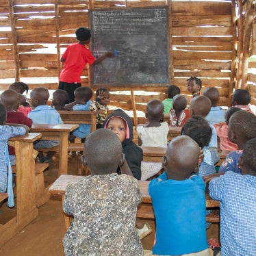 Young children sit in a rural classroom while a teacher writes on a blackboard