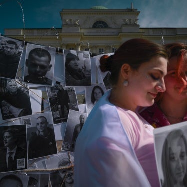 Two women hold a photo of a person in front of a fence of other photos of prisoners
