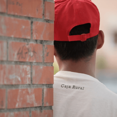 Closeup from behind of a teenager in a baseball cap and tshirt leaning against a brick column