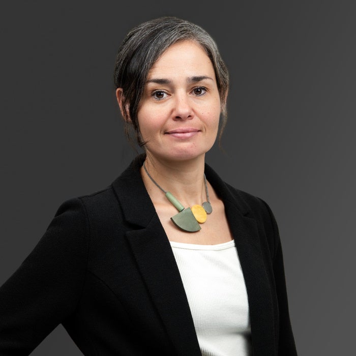  Studio headshot of a person with dark hair streaked with gray, wearing a black blazer, white top, and a necklace with green and gold geometric shapes. The person is facing the camera against a smooth dark gray background.